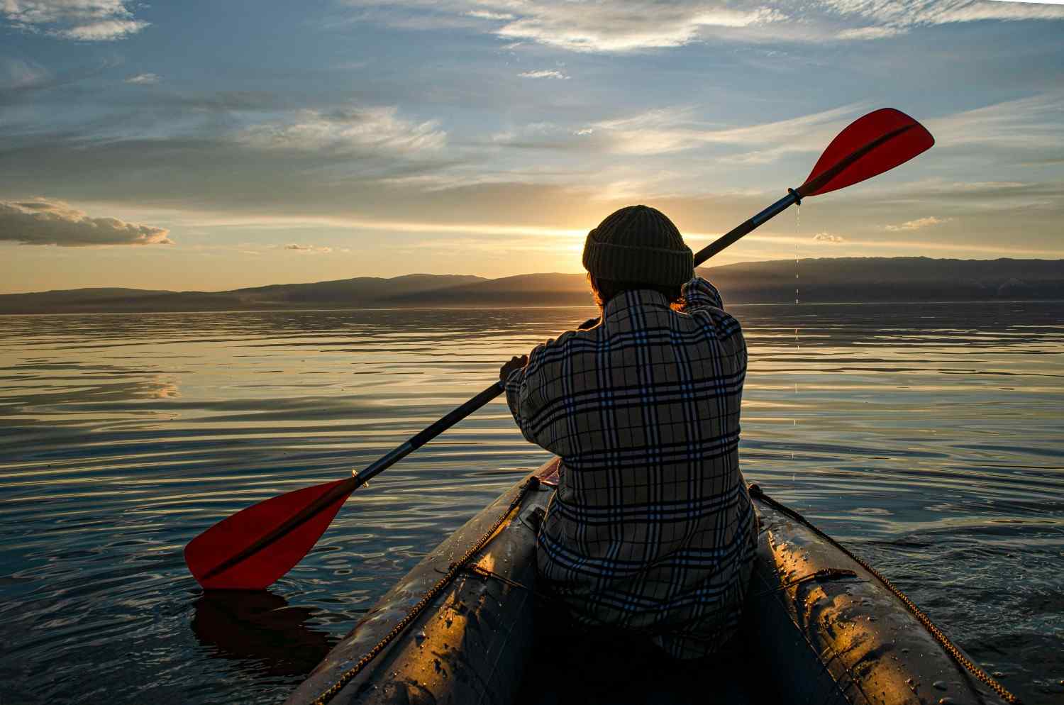 Back view of kayaker paddling with red paddle on calm lake at sunset