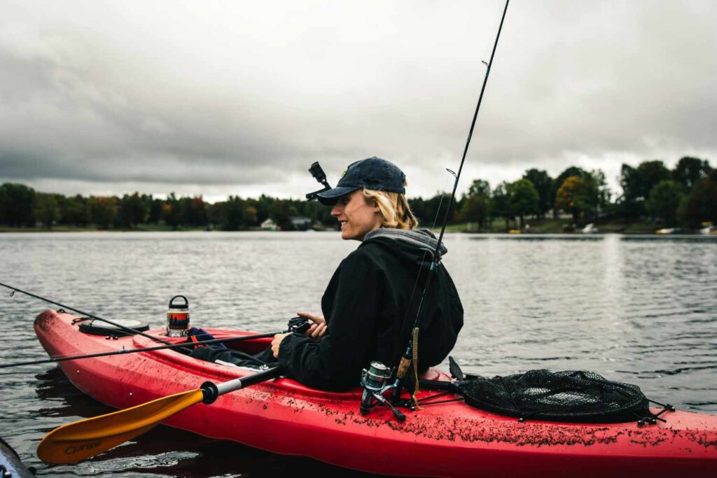Woman sitting in red fishing kayak with multiple fishing rods and paddle on lake