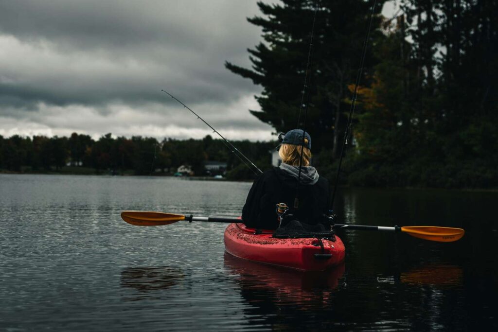 Woman sitting in wide red fishing kayak with fishing rods on calm lake near trees