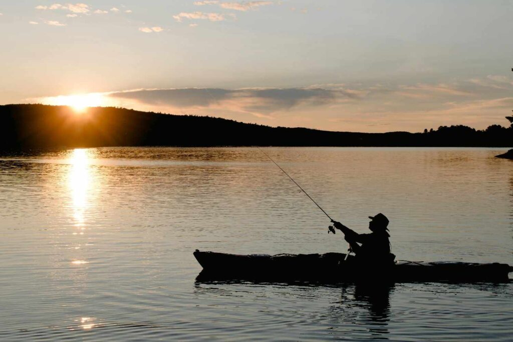 Silhouette of angler fishing from kayak on calm lake at sunset