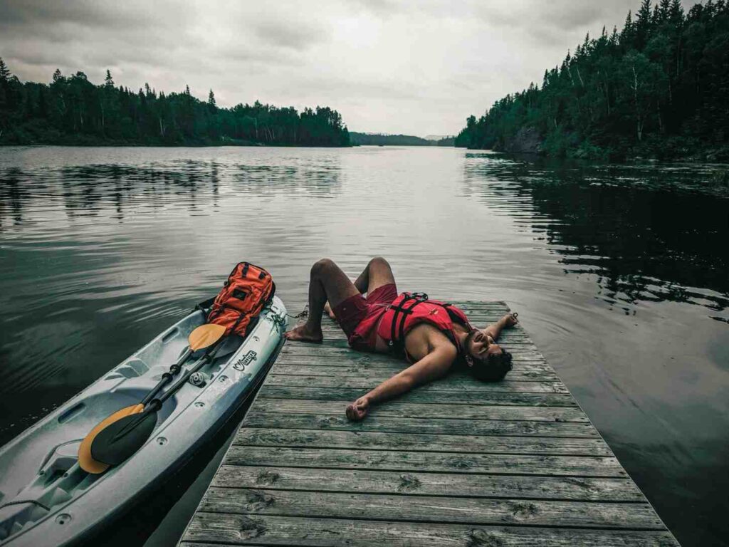 Exhausted kayaker lying on wooden dock next to kayak on calm lake