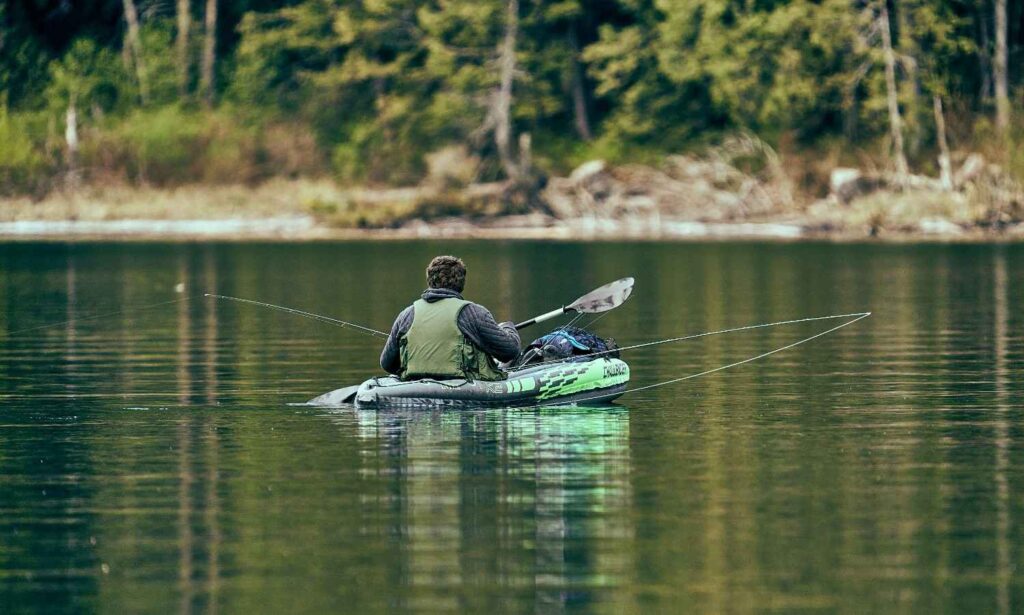 A man fishing from an inflatable Intex Challenger kayak on a calm green lake surrounded by forest with two fishing rods extended