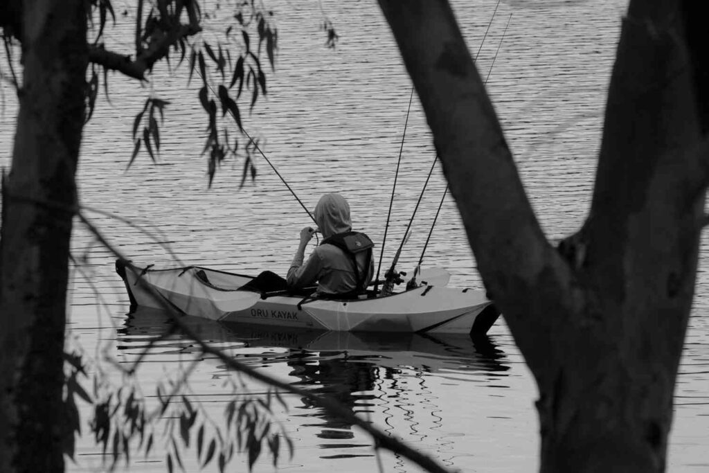Black and white photo of a person fishing from an Oru Kayak on still water surrounded by overhanging tree branches