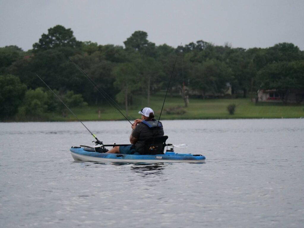 A man sitting in a blue Pelican fishing kayak on a calm lake with multiple fishing rods mounted and life vest on