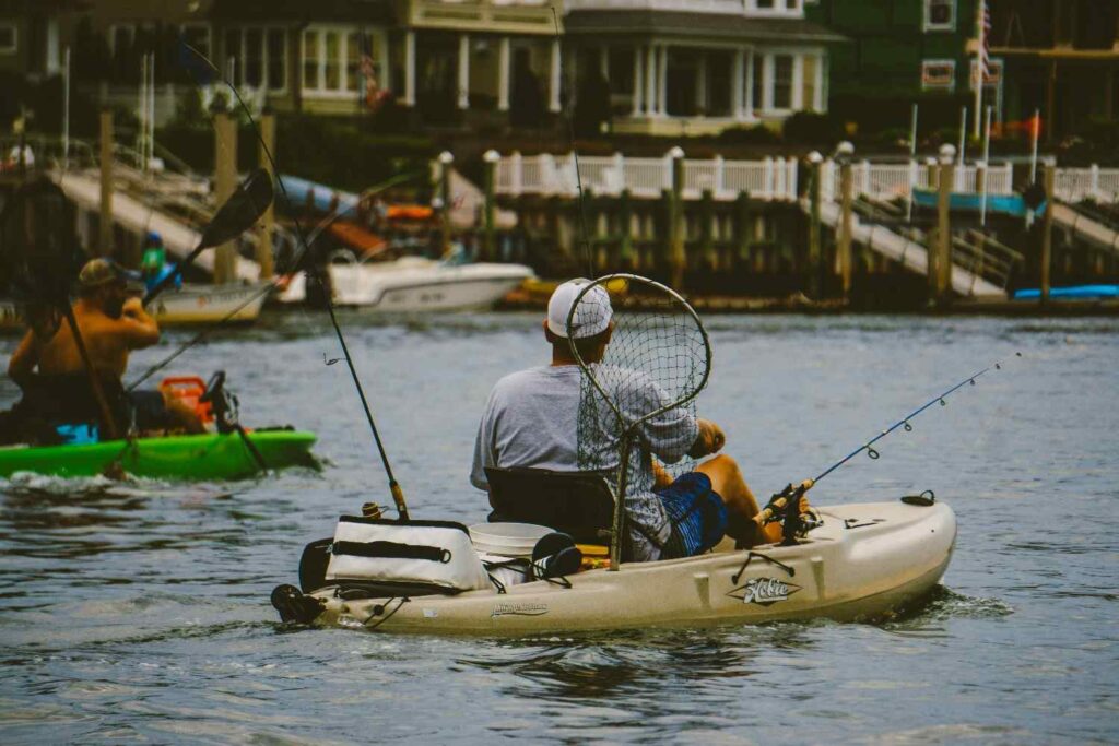A man sitting in a Hobie Mirage fishing kayak holding a landing net with fishing rods, cooler, and gear on board in a waterway near houses