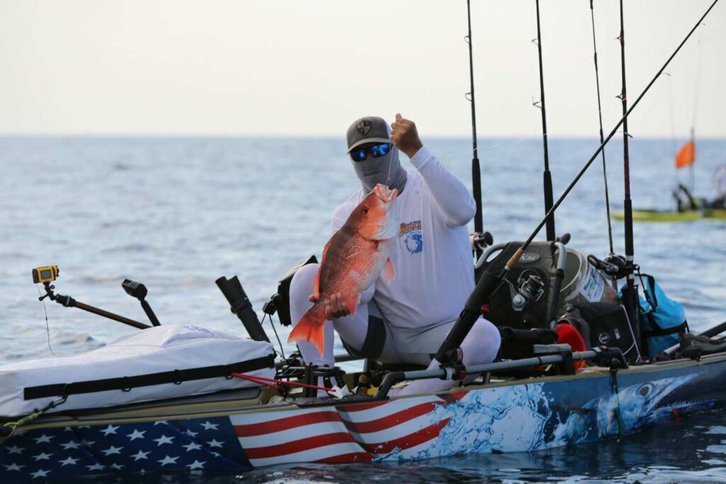 A man holding a large red snapper fish with thumbs up while sitting in a heavily rigged tournament fishing kayak on open water with multiple rods and camera mounts