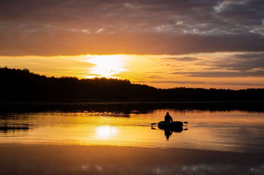  Silhouette of a lone angler sitting in a small boat on a perfectly calm lake with a dramatic golden sunset reflected on the water