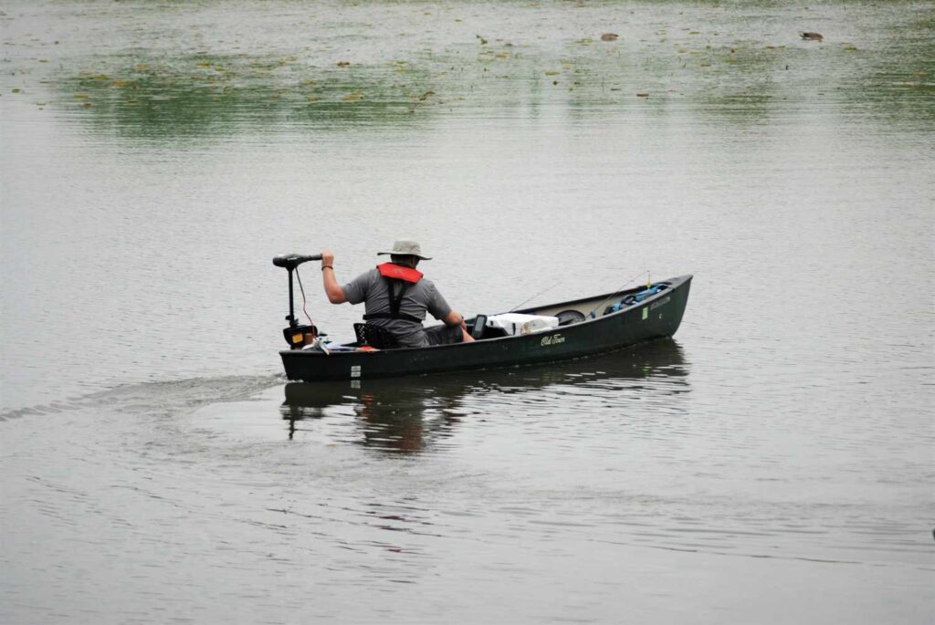A man operating a transom-mounted trolling motor on an Old Town fishing kayak on a calm lake with lily pads and fishing rods