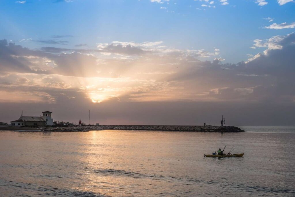 A person in a yellow fishing kayak paddling on calm water at sunrise with a lighthouse pier and dramatic sky in the background