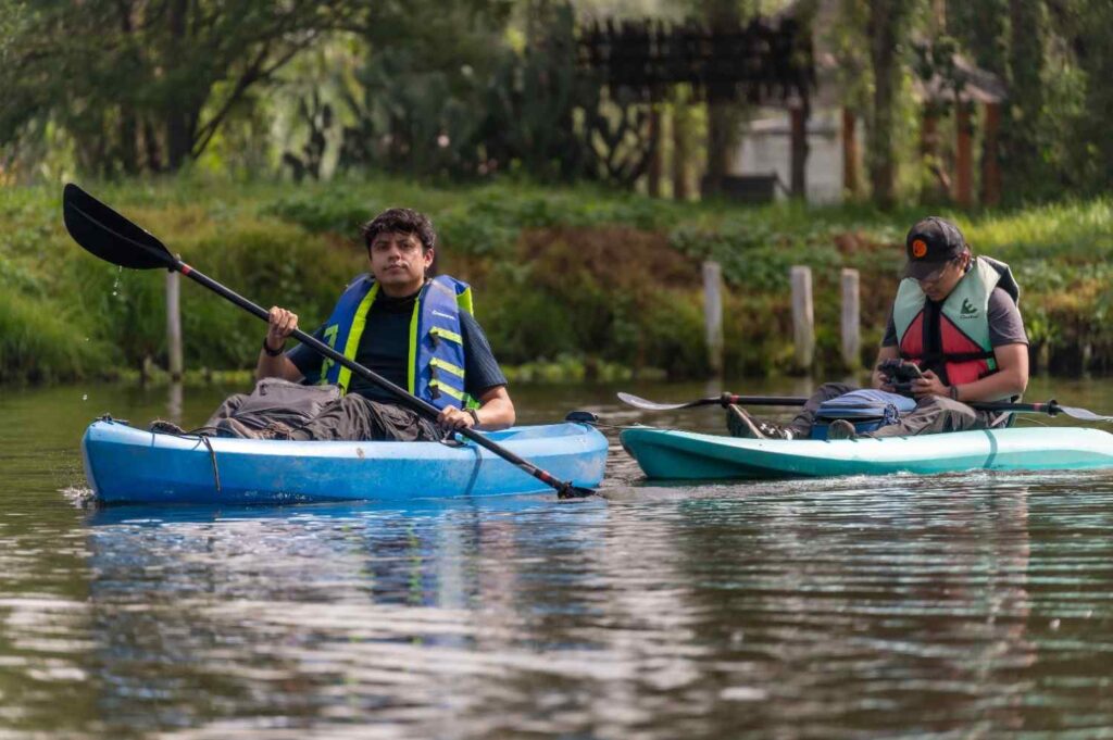 Two young men paddling solo sit-on-top kayaks on a calm river wearing life jackets surrounded by lush green vegetation