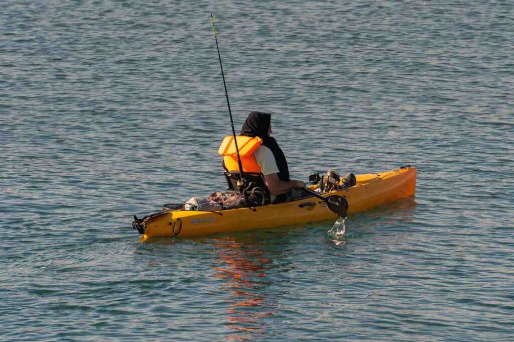  Man in orange life vest fishing from a yellow pedal kayak with rods deployed on warm open water