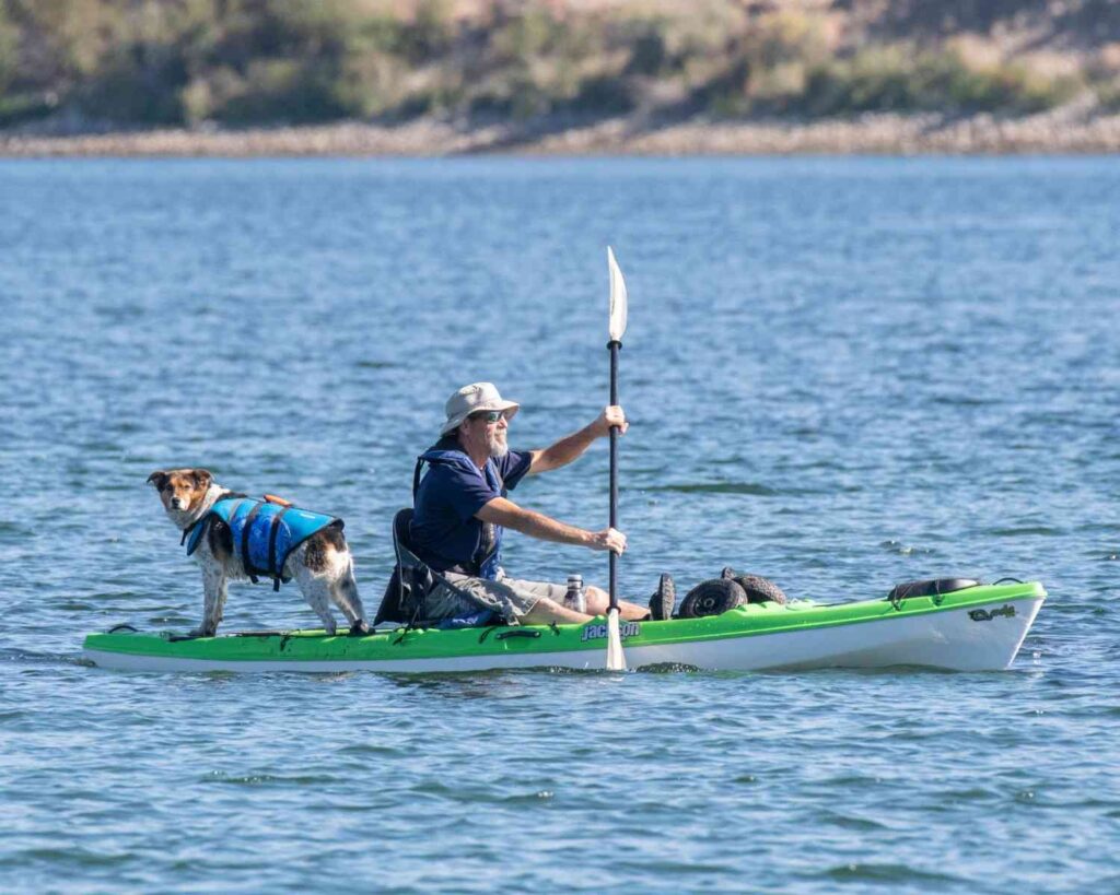 Bearded man in sun hat paddling a green Jackson fishing kayak with a dog standing on the front deck on open water