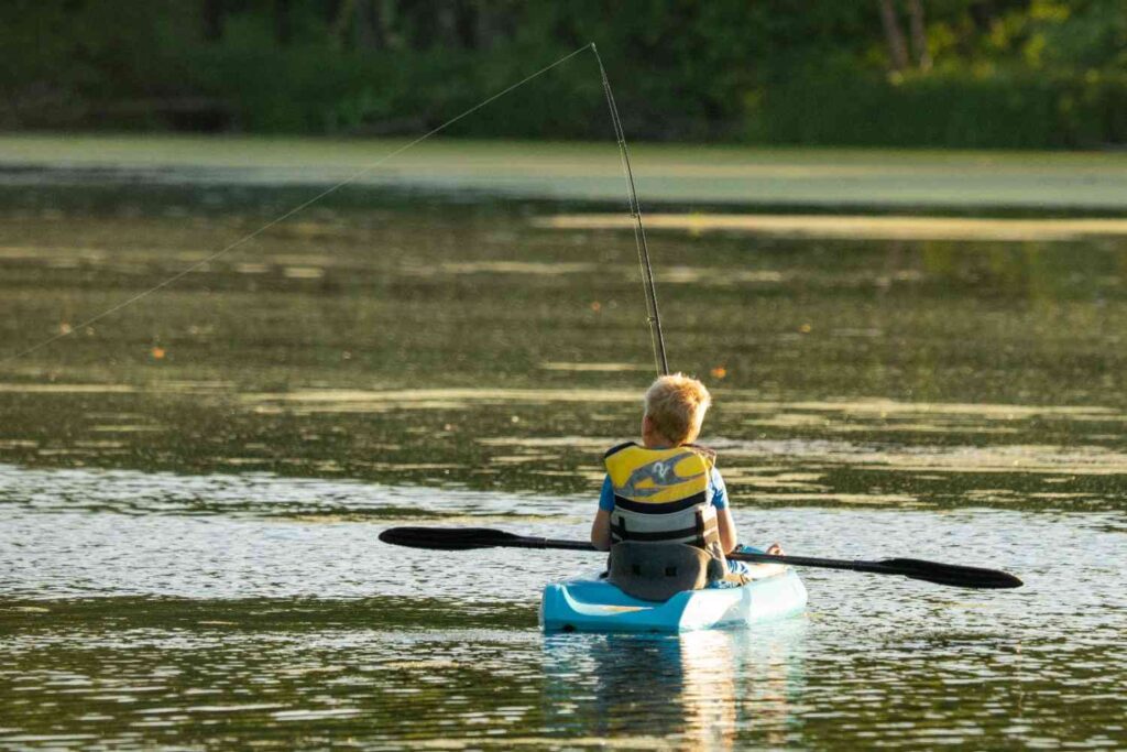 A young boy in a life jacket sitting in a blue kayak with a single fishing rod on a calm green freshwater lake at golden hour