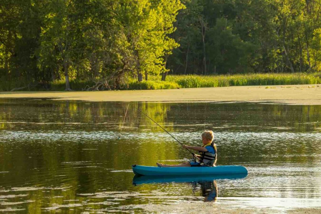A young boy in a life jacket fishing from a blue kayak near a weed edge on a calm freshwater lake surrounded by green trees at golden hour