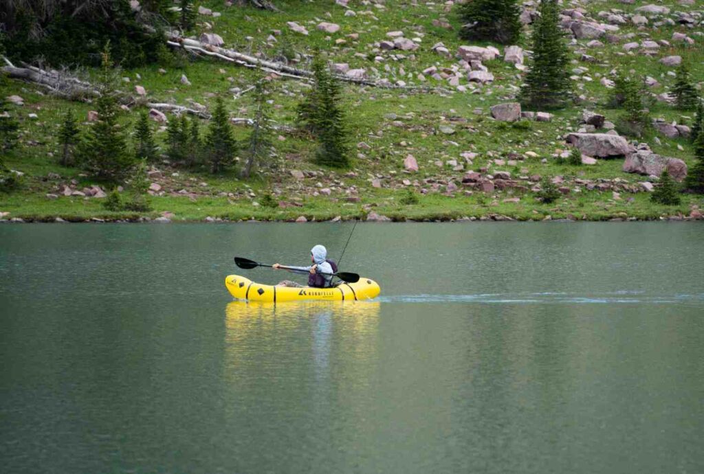 Person paddling a yellow inflatable kayak with fishing rod on a calm mountain lake surrounded by rocky slopes