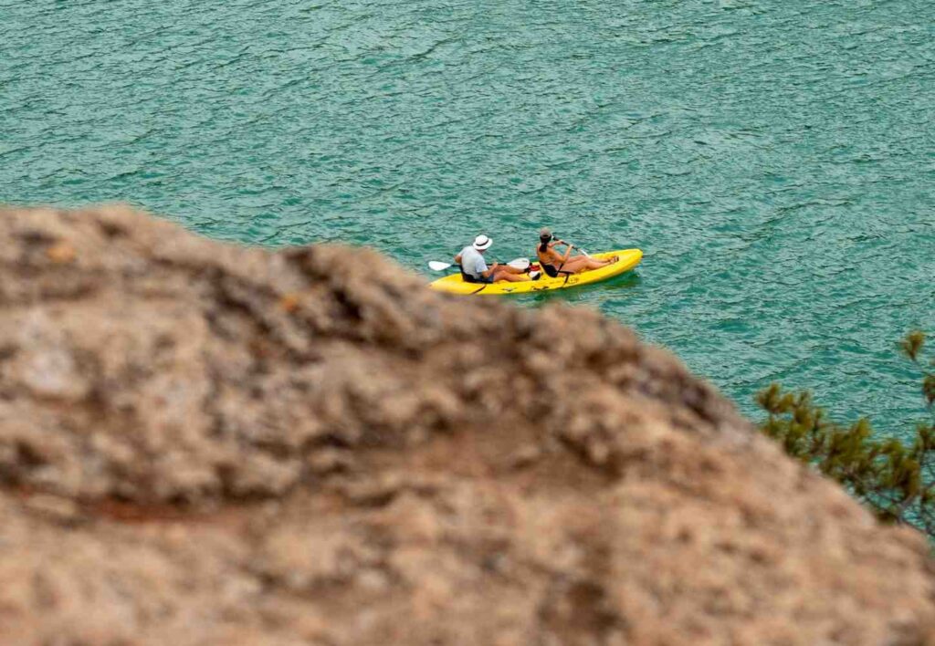 A man and woman paddling a yellow tandem kayak in a relaxed manner across calm turquoise water near rocky cliffs