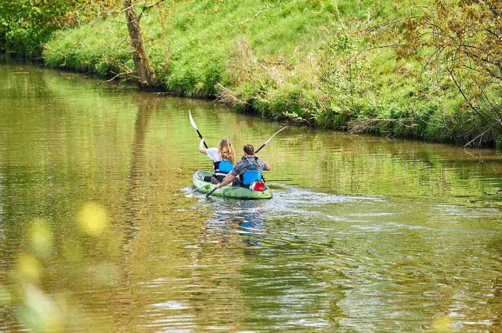 A man and woman wearing blue life jackets paddling a green tandem kayak together on a tranquil green river