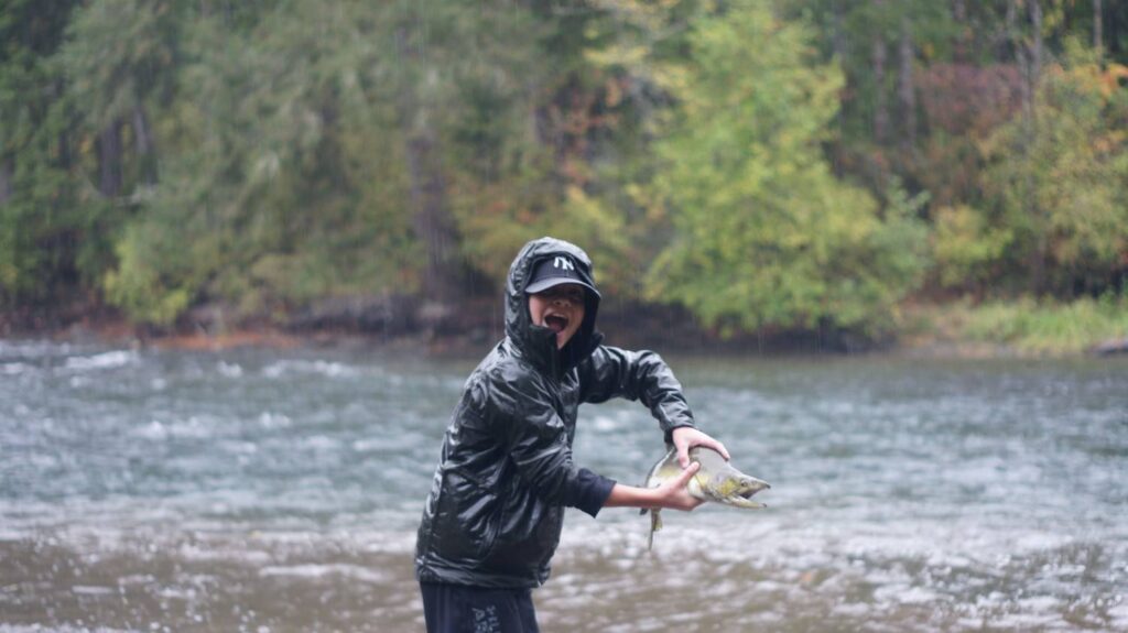 A young excited angler holding up a freshwater fish with a big open-mouthed smile while standing in a river in the rain