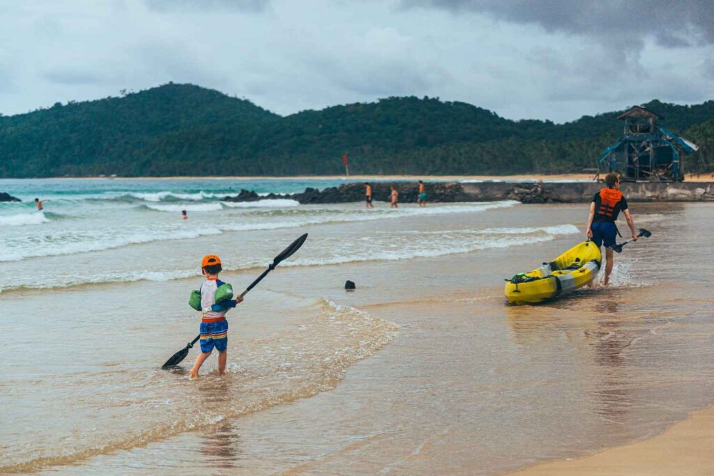 A child holding a paddle and an adult dragging a yellow kayak toward the water on a tropical beach