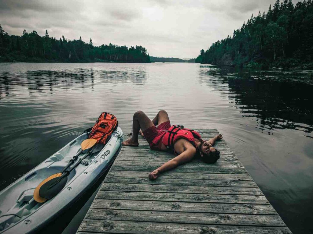 Tired man in life vest lying on wooden dock next to his kayak after a long fishing session on a dark forest lake