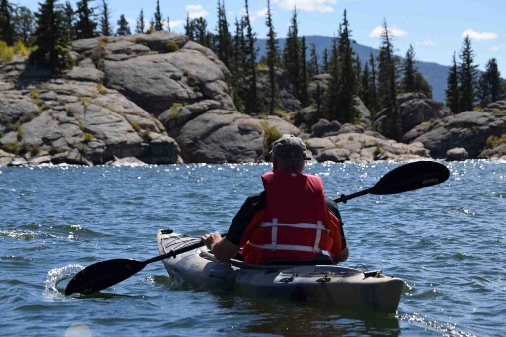 Back view of experienced kayaker in red life vest paddling confidently toward a rocky pine-lined mountain shoreline