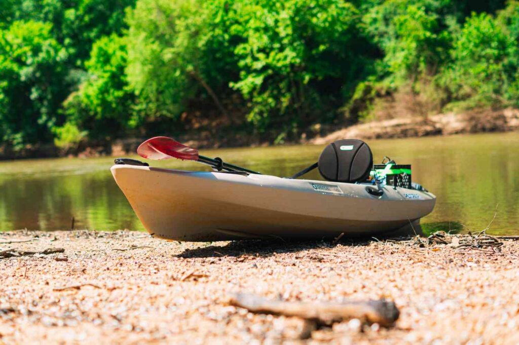 A tan fishing kayak resting on a sandy riverbank with a paddle, padded seat, and gear crate mounted at the back