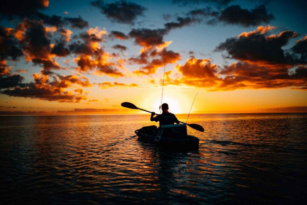 A silhouette of an angler paddling a fishing kayak with mounted rods toward a dramatic golden sunset on open water