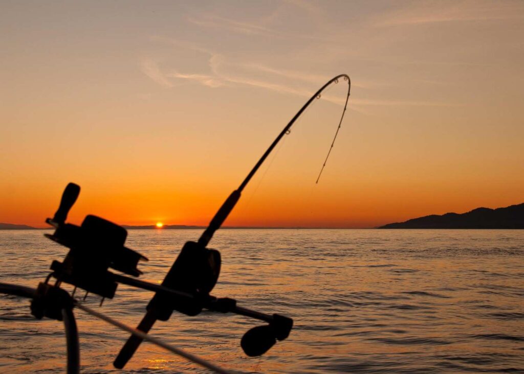 A fishing rod and reel silhouetted against a beautiful golden orange sunset over calm open water with mountain horizon