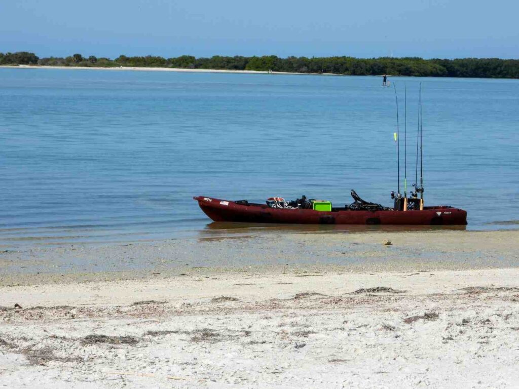 A dark red Maka fishing kayak resting on a sandy coastal beach fully loaded with multiple fishing rods, green gear crate, and equipment