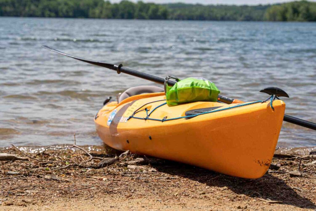 A green dry bag and paddle resting on top of an orange kayak parked on a lake shore