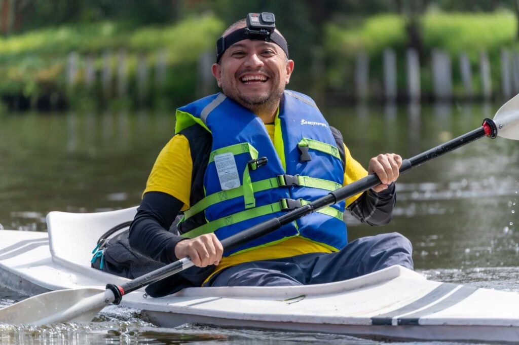 A smiling man wearing a blue life jacket and GoPro camera paddling a white kayak on a calm river