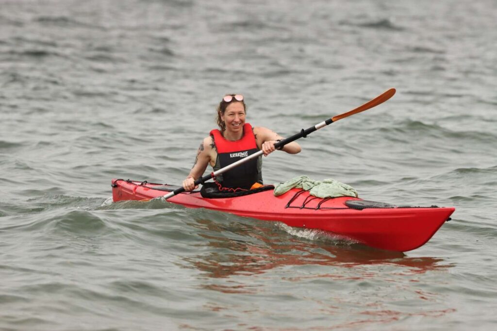 A smiling woman wearing a red and black life jacket paddling a red kayak confidently on open water