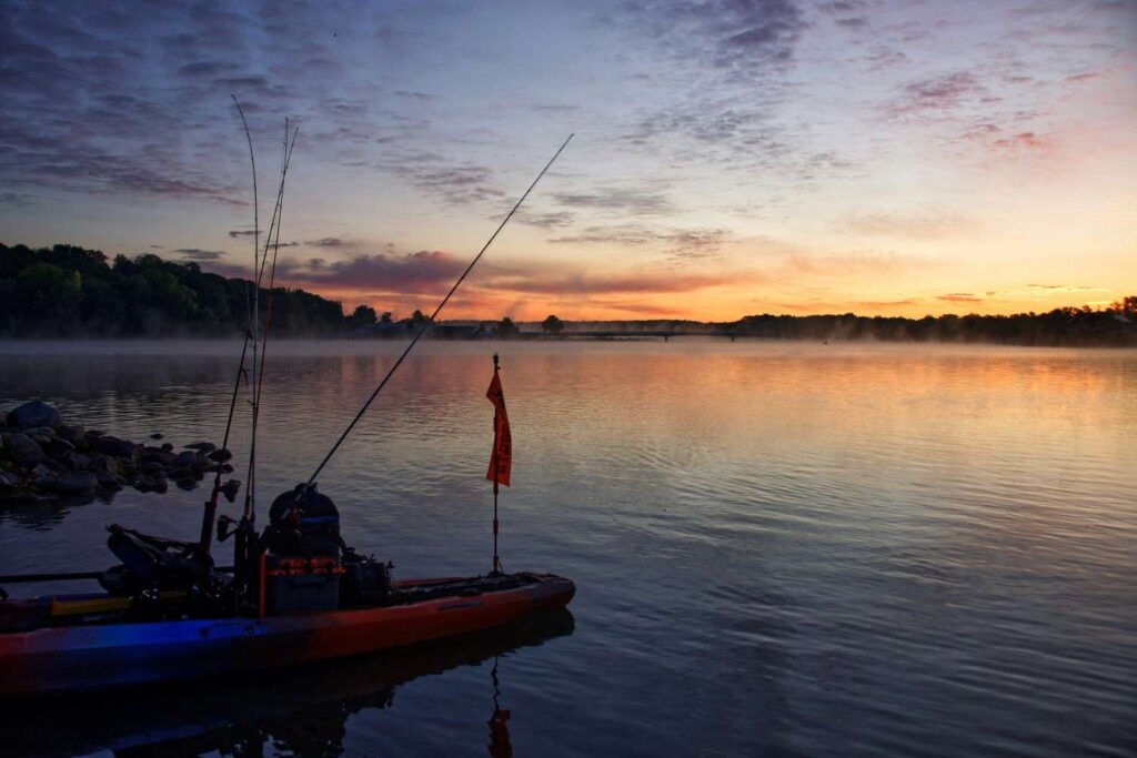 A heavily loaded fishing kayak with multiple rods, orange safety flag, and full gear setup resting at a rocky shoreline during a dramatic foggy sunrise
