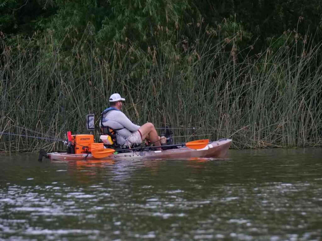 A man fishing from a fully loaded tan kayak near tall reeds with a fish finder, orange crate, cooler, and multiple fishing rods mounted