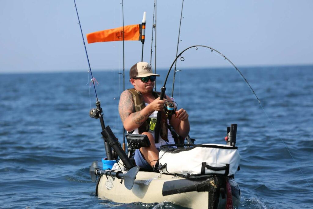 A man actively fishing from a heavily rigged kayak on open water with multiple rods, a fish finder, cooler, safety flag, and full gear setup