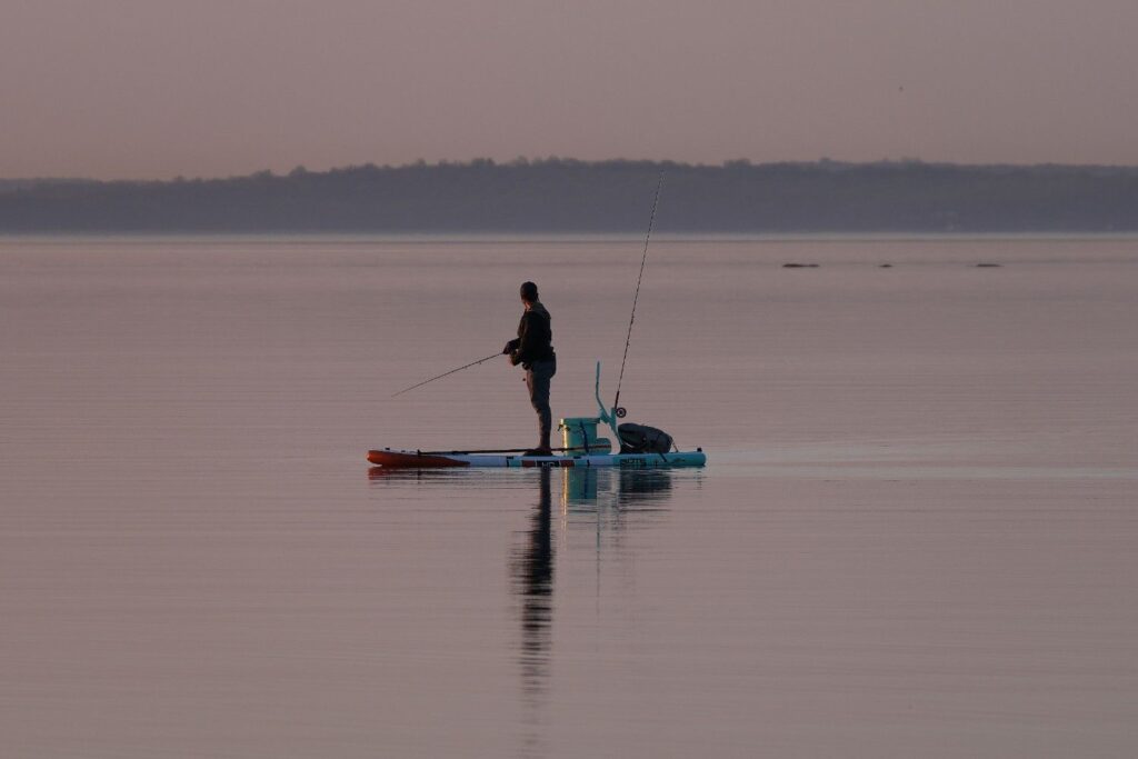 Man standing and fishing on a motorized pedal platform on a perfectly calm misty lake at dawn with rod deployed