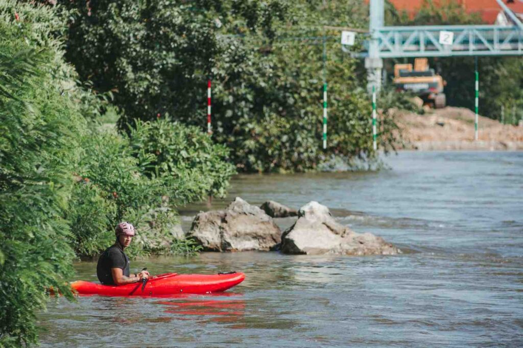 A man paddling a red inflatable kayak through rocky river water wearing a helmet and navigating around large boulders