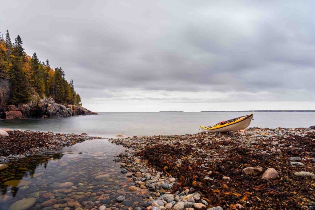 A yellow sea kayak resting on a rocky shoreline covered in pebbles and seaweed with calm water and rocky cliffs in the background