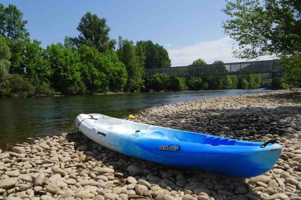A blue and white kayak resting on a rocky pebble shore beside a calm river with a bridge in the background