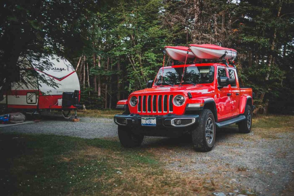 A red Jeep with a kayak strapped to the roof rack parked at a campsite surrounded by trees