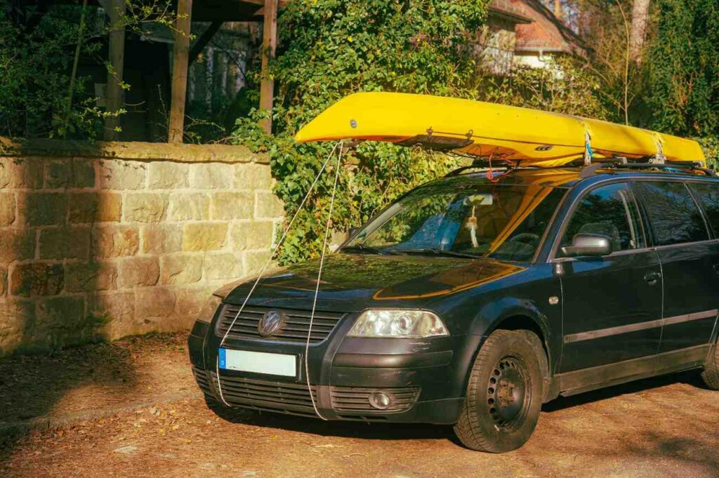 A yellow kayak secured with ropes on the roof rack of a black car parked outside, showing kayak transport setup