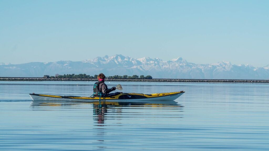A kayaker wearing a thermal layered jacket and neck gaiter paddling a yellow sea kayak on calm water with mountains in the background