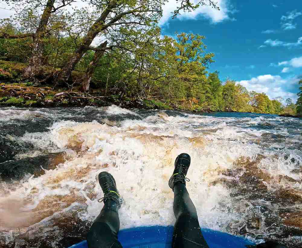First person view of a kayaker's black neoprene grip boots being splashed by cold whitewater on a river