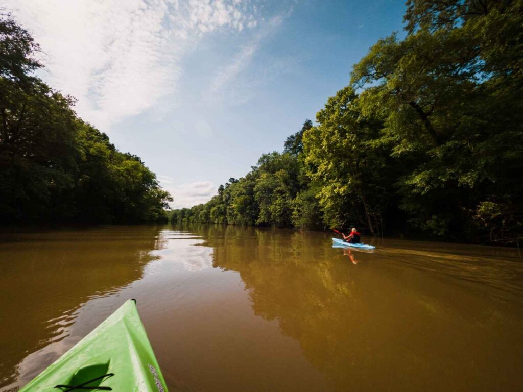 First-person view from a green kayak looking down a calm muddy river toward another kayaker paddling in the distance