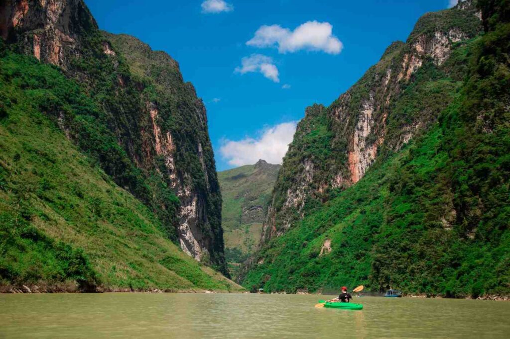 A kayaker paddling a green kayak positioned in a calm river flowing through a dramatic limestone canyon