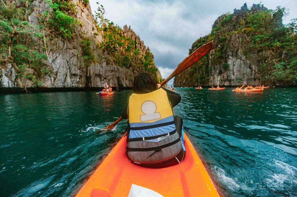 A kayaker in an orange kayak paddling between towering rocky cliffs on a scenic waterway