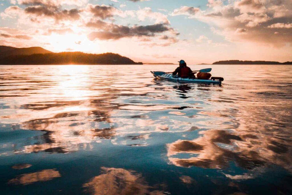 A kayaker silhouetted against a warm sunset paddling across calm reflective water with mountains in the background