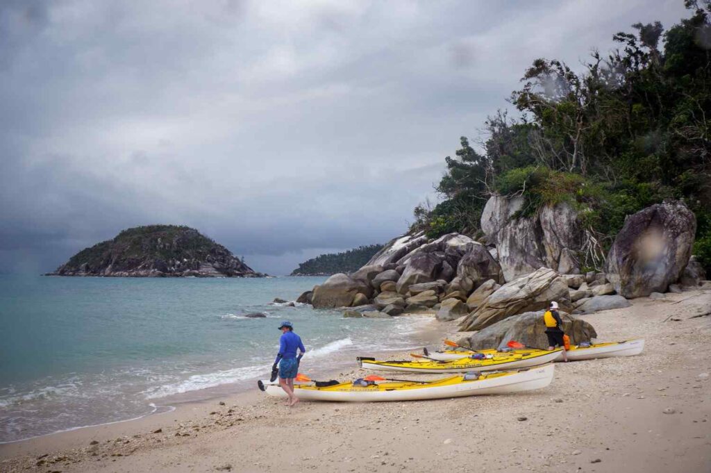 Two kayakers on a sandy beach with large rocks and boulders nearby, preparing their sea kayaks before launching into the water