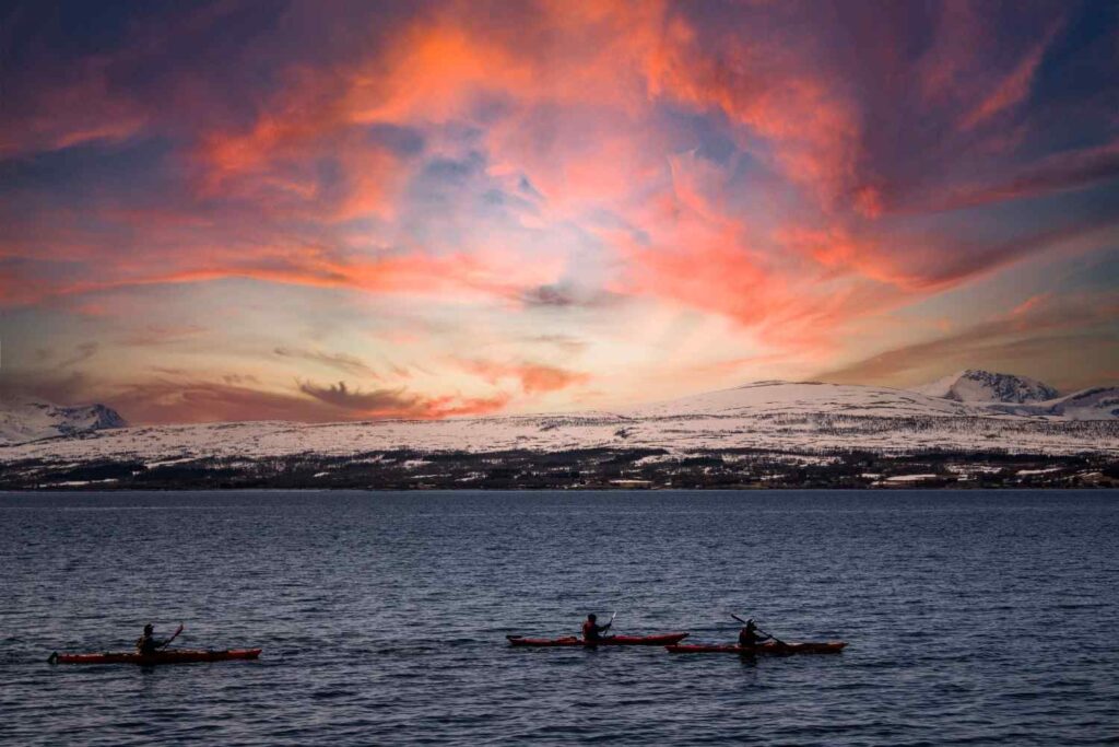 Silhouettes of three sea kayakers paddling on calm water beneath dramatic pink sunset clouds with snow-capped mountains in the background