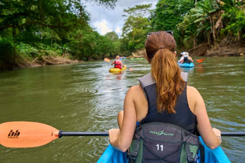 A kayaker paddling down a lush jungle river with other kayakers ahead, seen from a first-person perspective